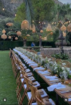 a long table is set up for an outdoor dinner party with hanging lights and greenery