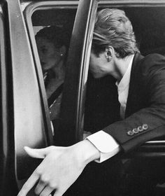 a man in a suit and tie leaning out the window of a car with his hand on the door handle