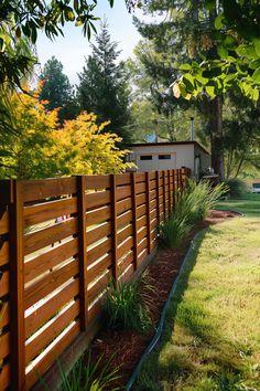 A wooden fence lines a garden path, with greenery and a variety of trees in the background on a sunny day. A small house is visible beyond the fence. Inexpensive Backyard Fence Ideas, Country Privacy Fence Ideas, Front Privacy Fence, Easy Cheap Fence Ideas, Vertical Privacy Fence Ideas, Wood Fence Ideas Backyards, Fence In Front Of House, Fence Around Tree, High Fence Ideas Privacy