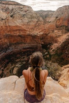 a woman sitting on top of a cliff looking at the canyon below her back view