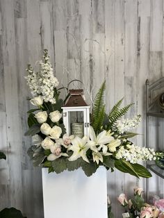 white flowers and greenery are arranged in a square vase with a lantern on top