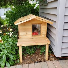 a cat sitting in a wooden house on the side of a building next to flowers