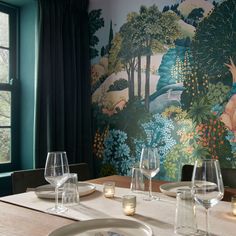 the table is set with wine glasses and place settings in front of a wall mural