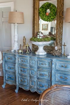 a blue dresser with an ornate mirror and wreath on it's top, next to two lamps