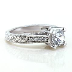 a white gold engagement ring with an intricate design on the band and a round diamond center stone