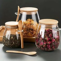 three glass jars filled with different types of flowers and herbs next to wooden spoons