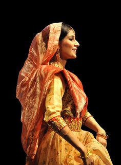 a woman in an orange and yellow dress sitting on top of a wooden bench next to a black background