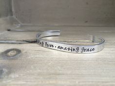 From the beloved Amazing Grace hymn Unending Love. Amazing Grace. is hand stamped on an aluminum bracelet. This bracelet measures 6” x 1/4” and can be formed to fit most wrist sizes. It looks great alone or layered with any of our other bracelets.  ~Metals Available~  Aluminum - We use 1100 Pure Food Safe Aluminum. This aluminum will not tarnish like sterling silver, or cause skin reactions like nickel, brass, or copper can. They are very lightweight and comfortable to wear. Aluminum is ... Silver Inspirational Bracelets For Valentine's Day, Inspirational Silver Bracelets For Valentine's Day, Inspirational Adjustable Hand Stamped Jewelry, Inspirational Silver Bracelet For Personalized Gift, Inspirational Hand Stamped Jewelry Bracelet, Inspirational Hand Stamped Bracelet Jewelry, Inspirational Hand Stamped Bracelet, Inspirational Hand-stamped Bracelet Jewelry, Inspirational Adjustable Silver Name Bracelet