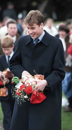 a man in a long black coat is holding flowers and presents while other people look on