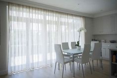 a dining room table with white chairs next to a large window covered in sheer curtains