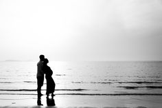 a man and woman standing on top of a beach next to the ocean with their arms around each other