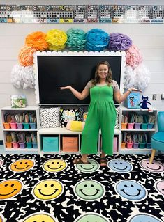 a woman standing in front of a blackboard with smiley faces on it and colorful decorations behind her