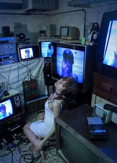 a woman sitting on the floor in front of televisions and other electronic equipment,