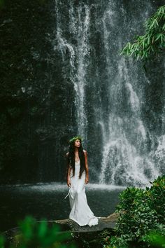 a woman standing in front of a waterfall wearing a white dress and holding a flower crown