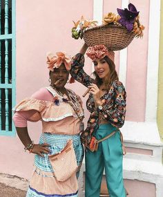 two women standing next to each other in front of a pink building with baskets on their heads