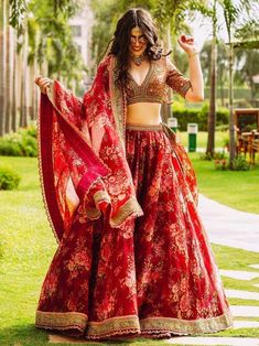 a woman in a red and gold lehenga dancing on the grass with palm trees behind her