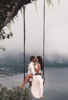 two people are sitting on a swing in front of the water and trees, one person is kissing the other