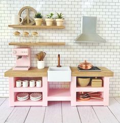 a toy kitchen with pink cabinets and white tiles on the wall behind it is an oven, sink, stovetop, and shelves filled with pots and pans