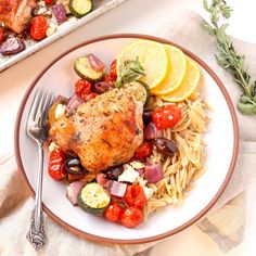 a white plate topped with chicken, pasta and veggies next to a tray of lemon wedges