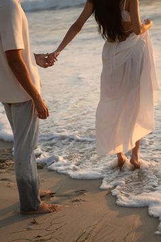 a man and woman holding hands while standing on the beach at sunset with waves coming in