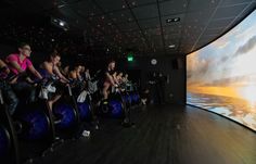 there are many people riding stationary bikes in front of a large screen at the same time