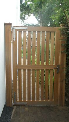 an open wooden gate in front of a white wall and brick walkway with trees on either side