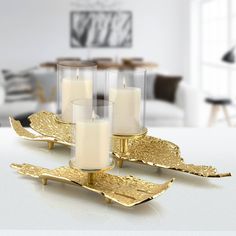 three candles sitting on top of gold leaf shaped trays in front of a white table