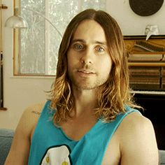 a man with long hair sitting on a couch in front of a piano and looking at the camera