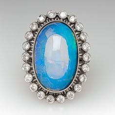 This gorgeous ring is centered with one (1) oval cabochon cut natural crystal opal and set into a bezel setting. The opal is bordered with twenty-two (22), bezel set, round brilliant cut diamonds, The top face of the shank is accented with three (3), bead set, round brilliant cut diamonds on each side. The gemstones are bordered with milgrain edging. The ring measures 29.1mm at the top, rises 10.6mm above the finger, tapering to 1.8mm wide and 1.1mm thick at the base of the shank. This ring is currently a size 6. Oval Cabochon Opal Ring Fine Jewelry, Fine Jewelry Oval Cabochon Opal Ring, Oval Cabochon Opal Ring In Fine Jewelry, Oval Opal Ring With Bezel Setting For Formal Occasions, Oval Cabochon Diamond Opal Ring, Opal Diamond Ring Oval Cabochon, Diamond Opal Ring Oval Cabochon, Oval Cabochon White Gold Opal Ring, Oval Cabochon Opal Ring In White Gold