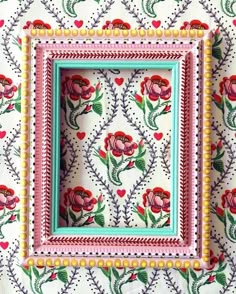 an ornate frame with flowers and hearts painted on the wall next to a flowered tablecloth