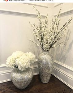 two silver vases with white flowers in them sitting on a wooden table next to a wall