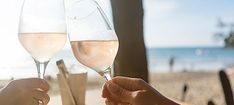 two people toasting with wine glasses on a table at the beach in front of the ocean