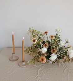 two candles are sitting next to a vase with flowers in it on a white table cloth