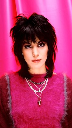 a woman with black hair wearing a pink sweater and necklaces, posing for the camera