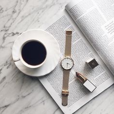 a cup of coffee, watch and lipstick sitting on top of an open book