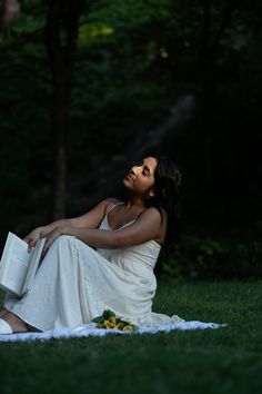 a woman is sitting on the grass reading a book and looking up into the sky