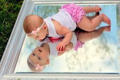 a baby laying on the ground in front of a mirror with grass and flowers around it