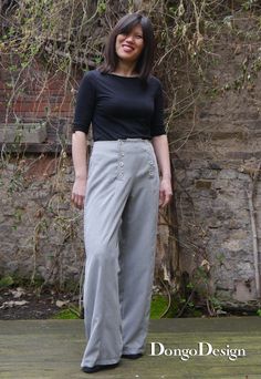 a woman standing in front of a brick wall wearing grey pants and black top with buttons on the side