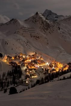 a town is lit up at night in the mountains with snow on the ground and trees around it