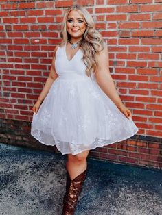 a woman standing in front of a brick wall wearing a white dress and cowboy boots