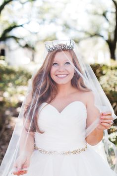 a woman in a wedding dress and tiara smiles as she holds her veil over her head