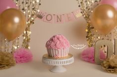 a pink cupcake sitting on top of a white cake plate next to gold balloons