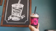 a person holding up a drink in front of a chalkboard with an advertisement on it