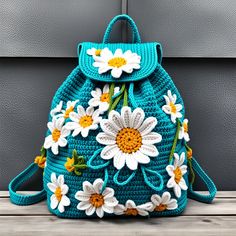 a crocheted backpack with white and yellow daisies on the front, attached to a wooden bench