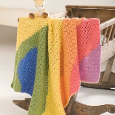 a crocheted blanket with a teddy bear sitting on it's back in front of a rocking chair