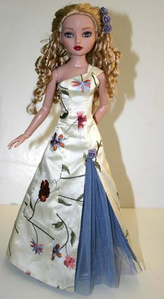 a doll with blonde hair wearing a white dress and blue flowered skirt, standing in front of a white wall