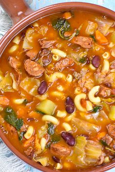 a bowl filled with pasta and sausage soup on top of a blue cloth next to a wooden spoon