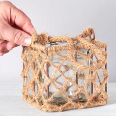 a hand holding a small glass jar with rope around the top and bottom, in front of a white background