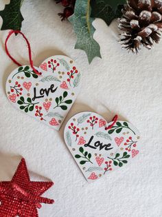 two heart shaped ornaments sitting on top of a white surface next to pine cones and evergreen leaves