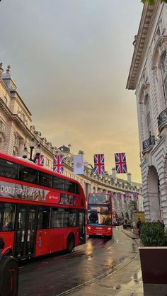 two red double decker buses driving down a street next to tall buildings with flags on them